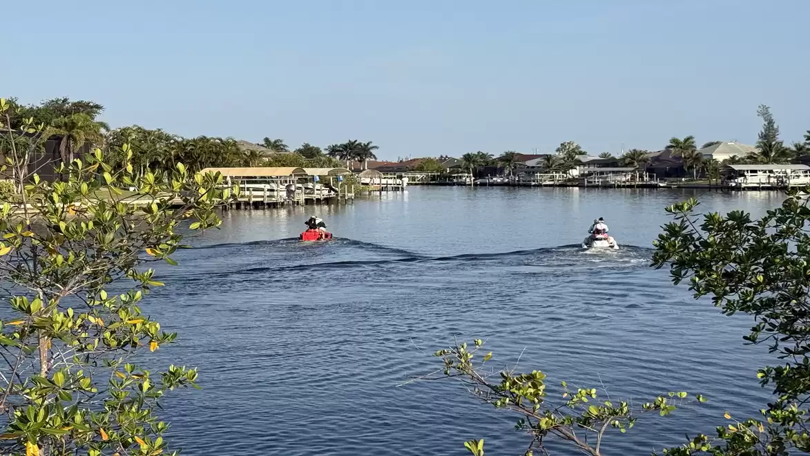Two people riding jet skis on a calm lake surrounded by trees and houses under a clear sky.