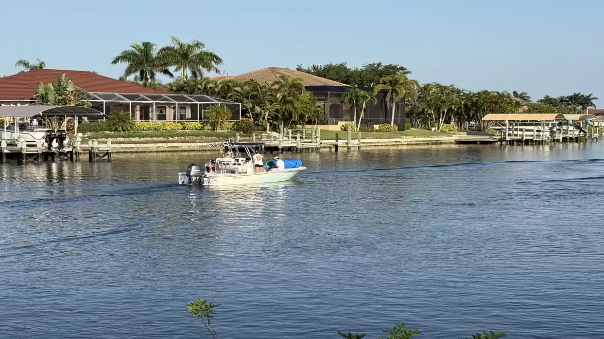 A motorized boat sails on a calm canal past modern houses with palm trees in the background.