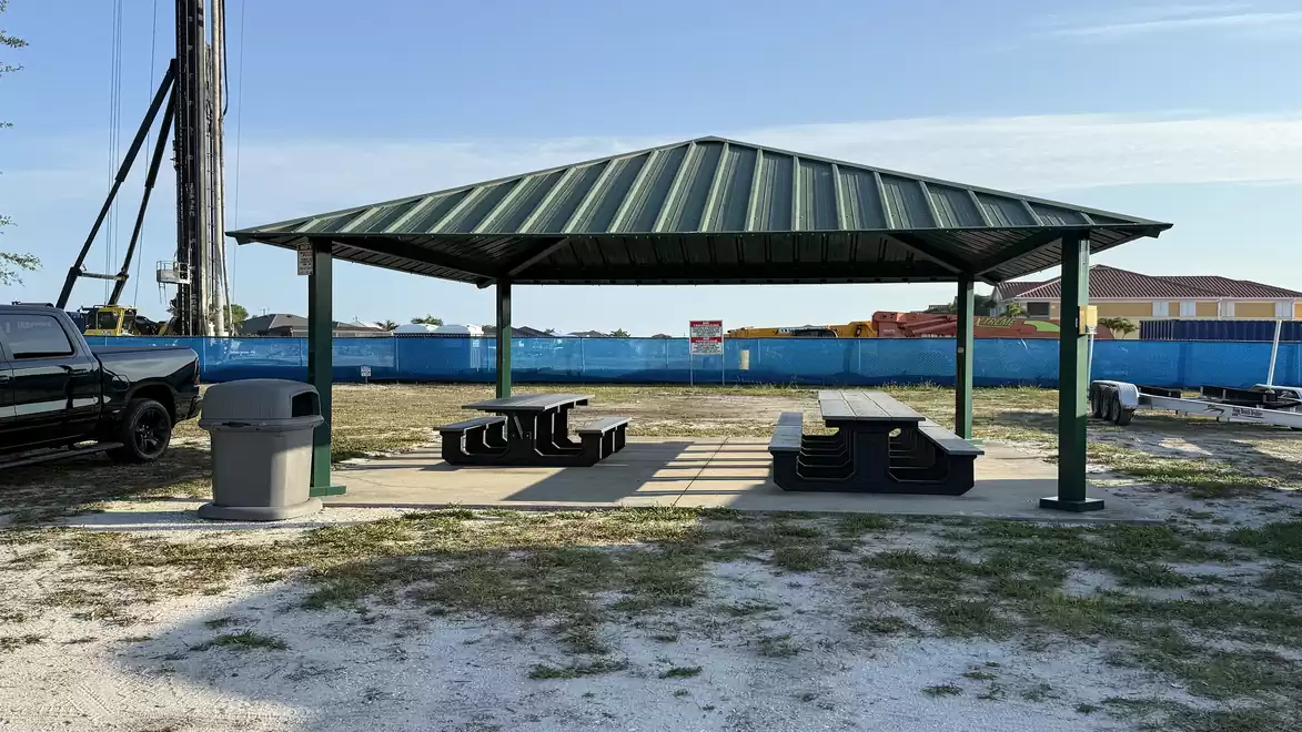 A pavilion with a green metal roof, with picnic tables underneath on a concrete surface. A black pickup truck and a trash can are on the left, with a blue construction fence in the background.