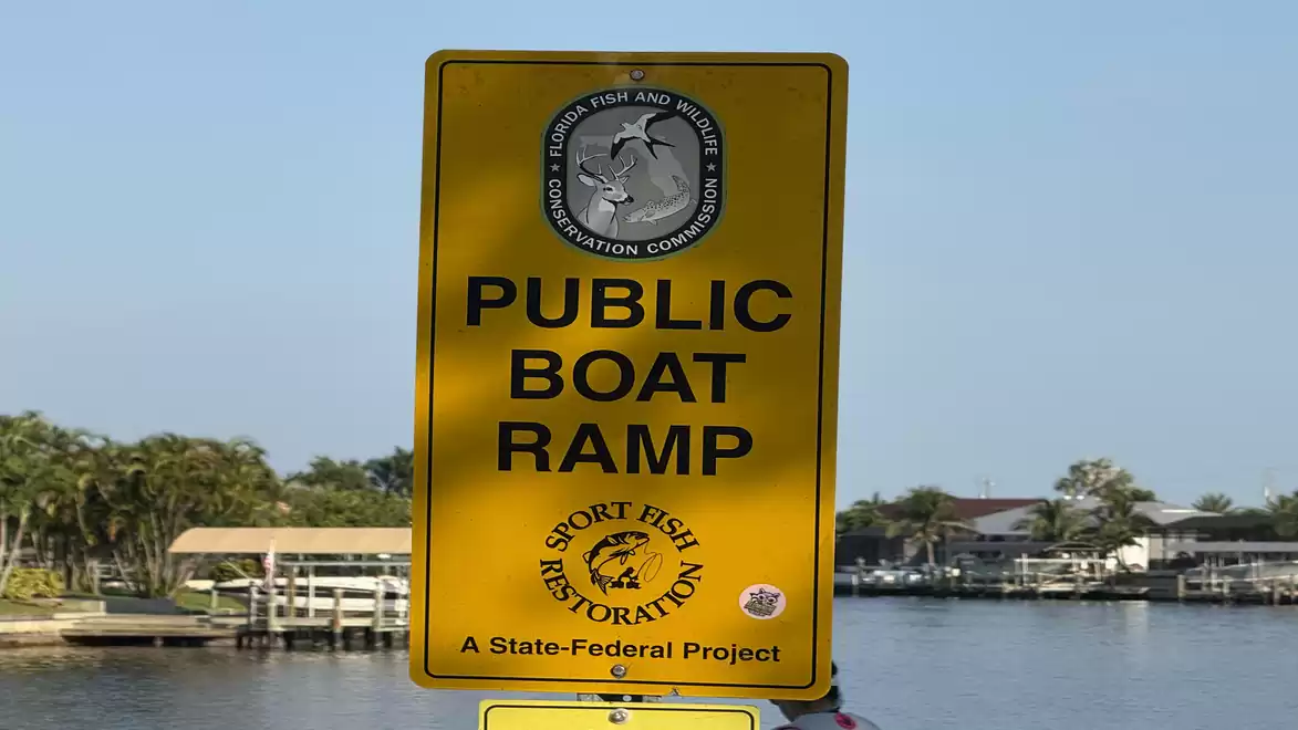 A yellow sign reading 'Public Boat Ramp' in front of a water background with houses and trees on the shore.