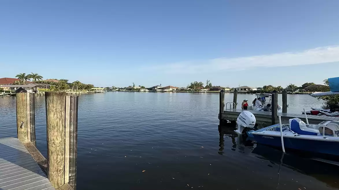 A serene harbor with several boats under a clear blue sky. In the foreground are wooden posts and a boat with a blue roof.