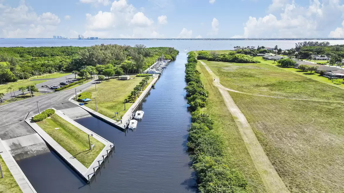 Aerial view of a canal with boats, surrounded by green fields and trees, with the sea and blue sky in the background.