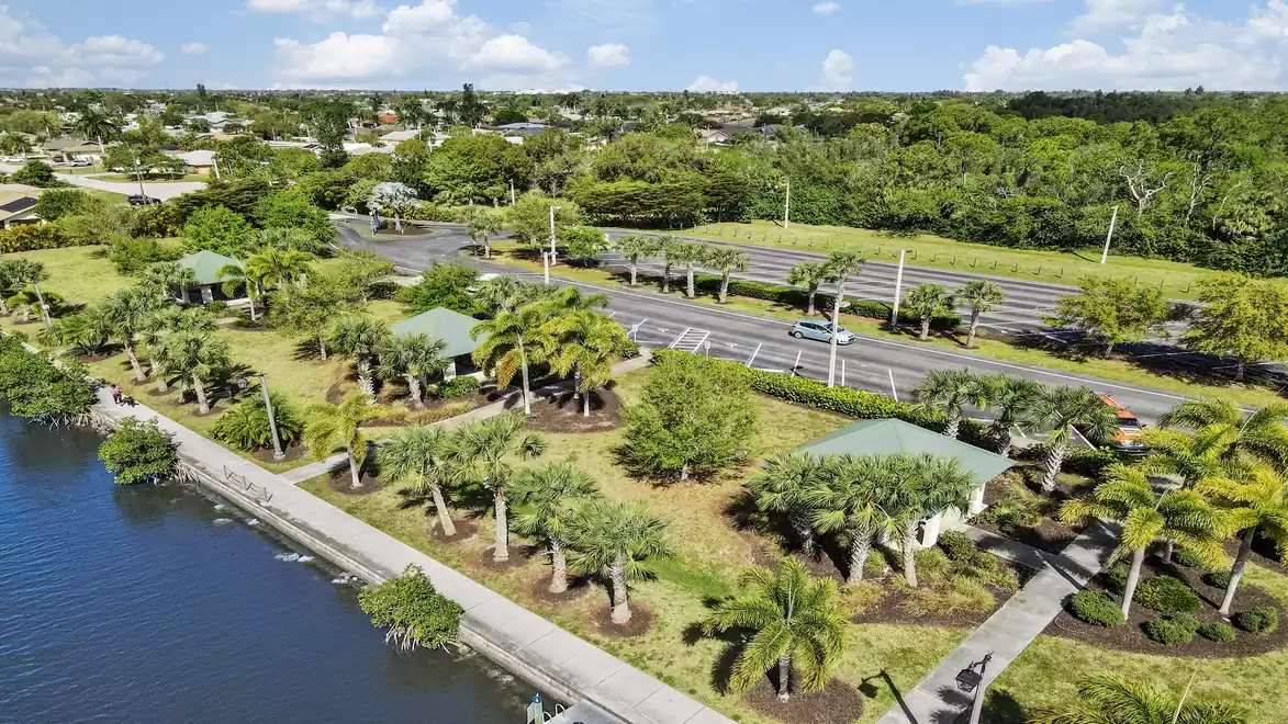 Aerial view of a green park with palm trees located by a body of water. In the background, a road runs through lush greenery under a clear blue sky.