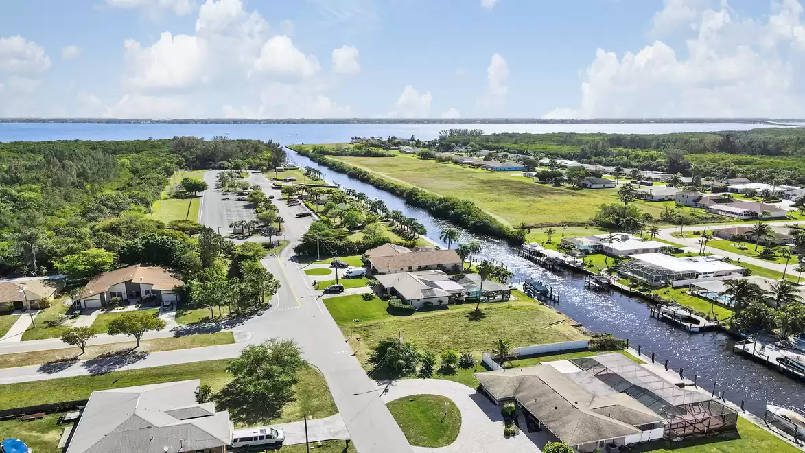 Aerial view of a residential area with a canal winding through the landscape. Several houses with well-maintained gardens are arranged along the streets and canals. In the background, a dense wooded area is visible, while the sky is partly cloudy.