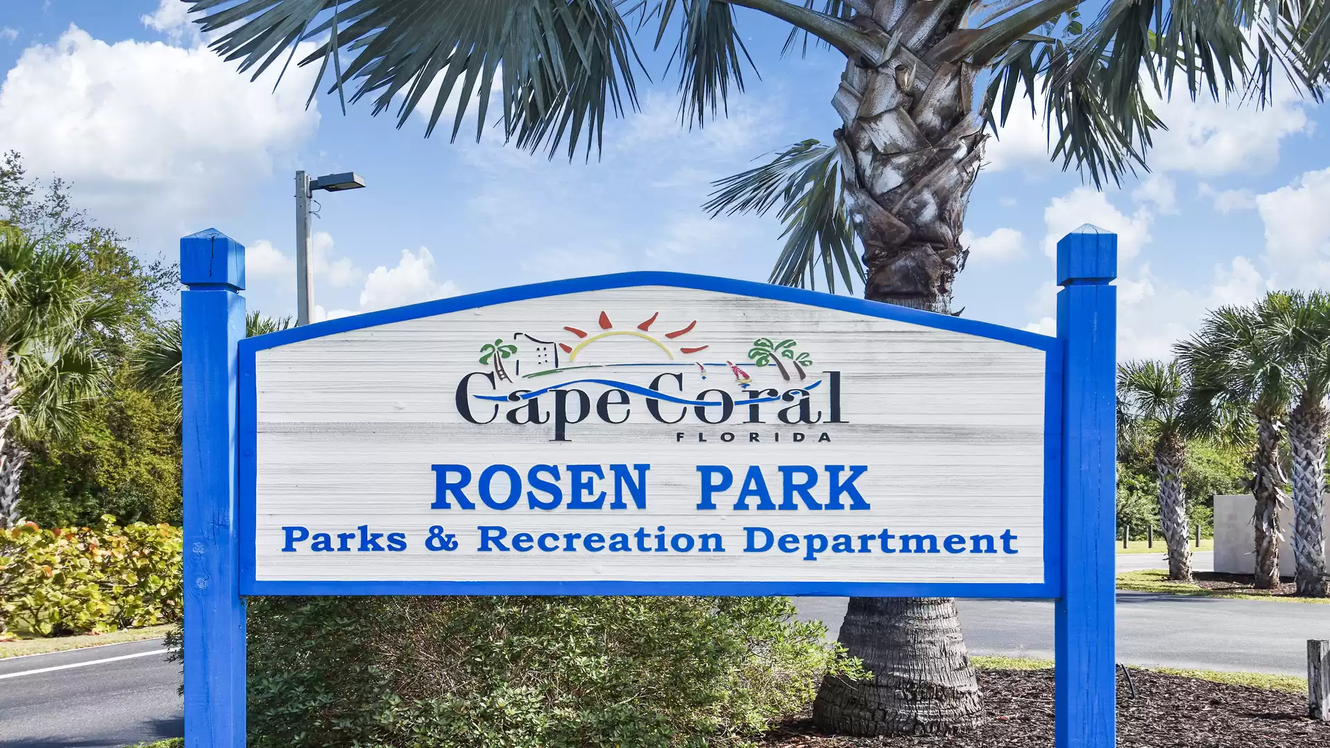 A sign marks Rosen Park in Cape Coral, Florida. It is surrounded by palm trees and displays the text 'Parks & Recreation Department'.