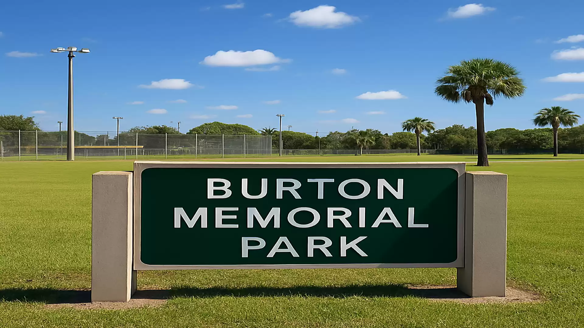 A green sign at Burton Memorial Park with a well-maintained lawn and palm trees in the background under a clear blue sky.