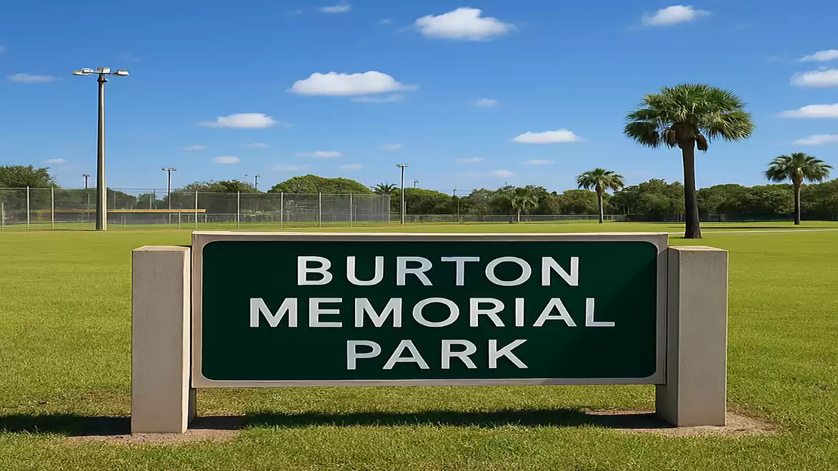A green sign at Burton Memorial Park with a well-maintained lawn and palm trees in the background under a clear blue sky.