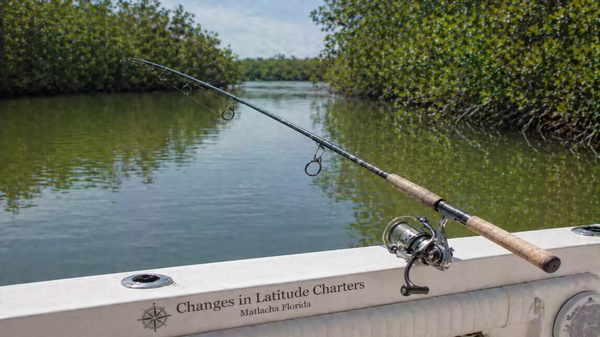 A fishing rod rests on the railing of a boat anchored in a calm lagoon surrounded by mangroves in Florida.