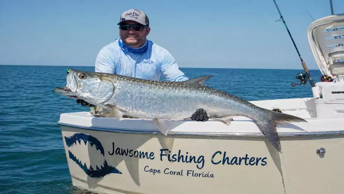 An angler holds a large fish on a boat in the ocean. The boat reads 'Jawsome Fishing Charters, Cape Coral Florida'.