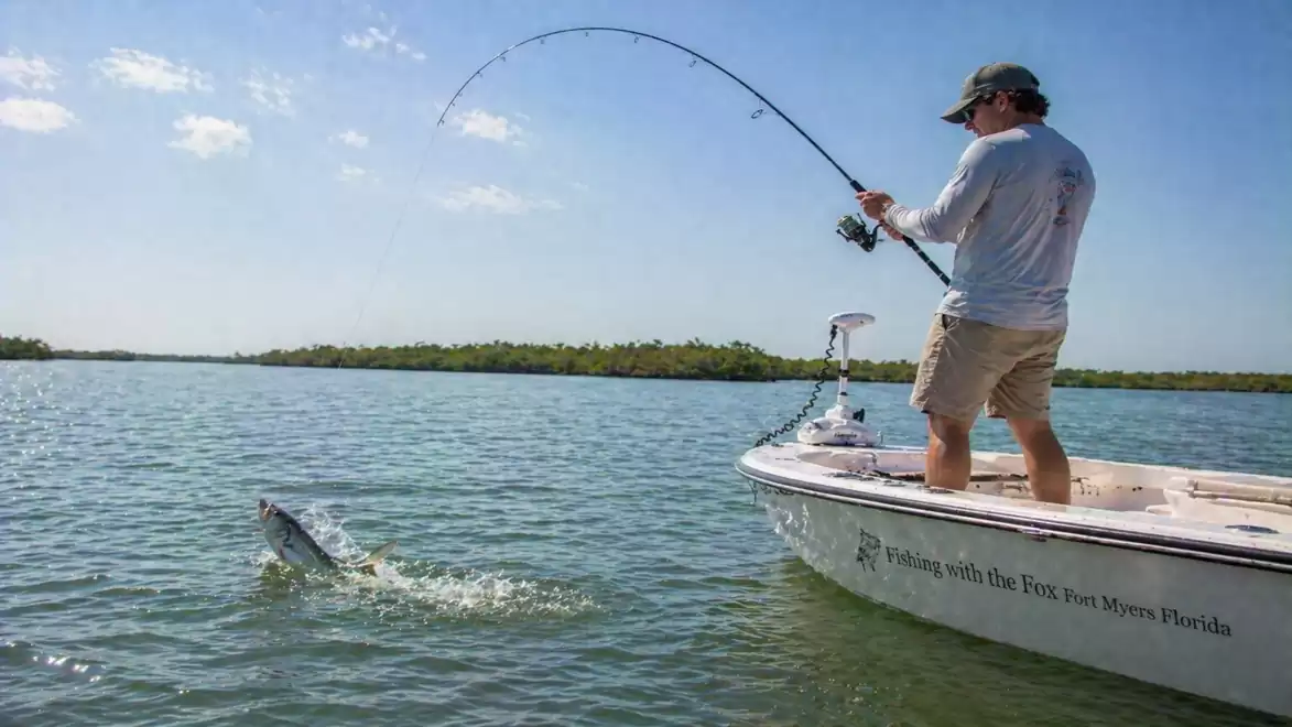 A man is fishing on a boat in a sunny body of water. He is reeling in a large fish.