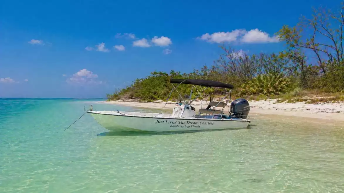 A small motorboat with a sunshade is docked at the clear turquoise waters of a tropical island. Trees and blue sky are visible in the background.