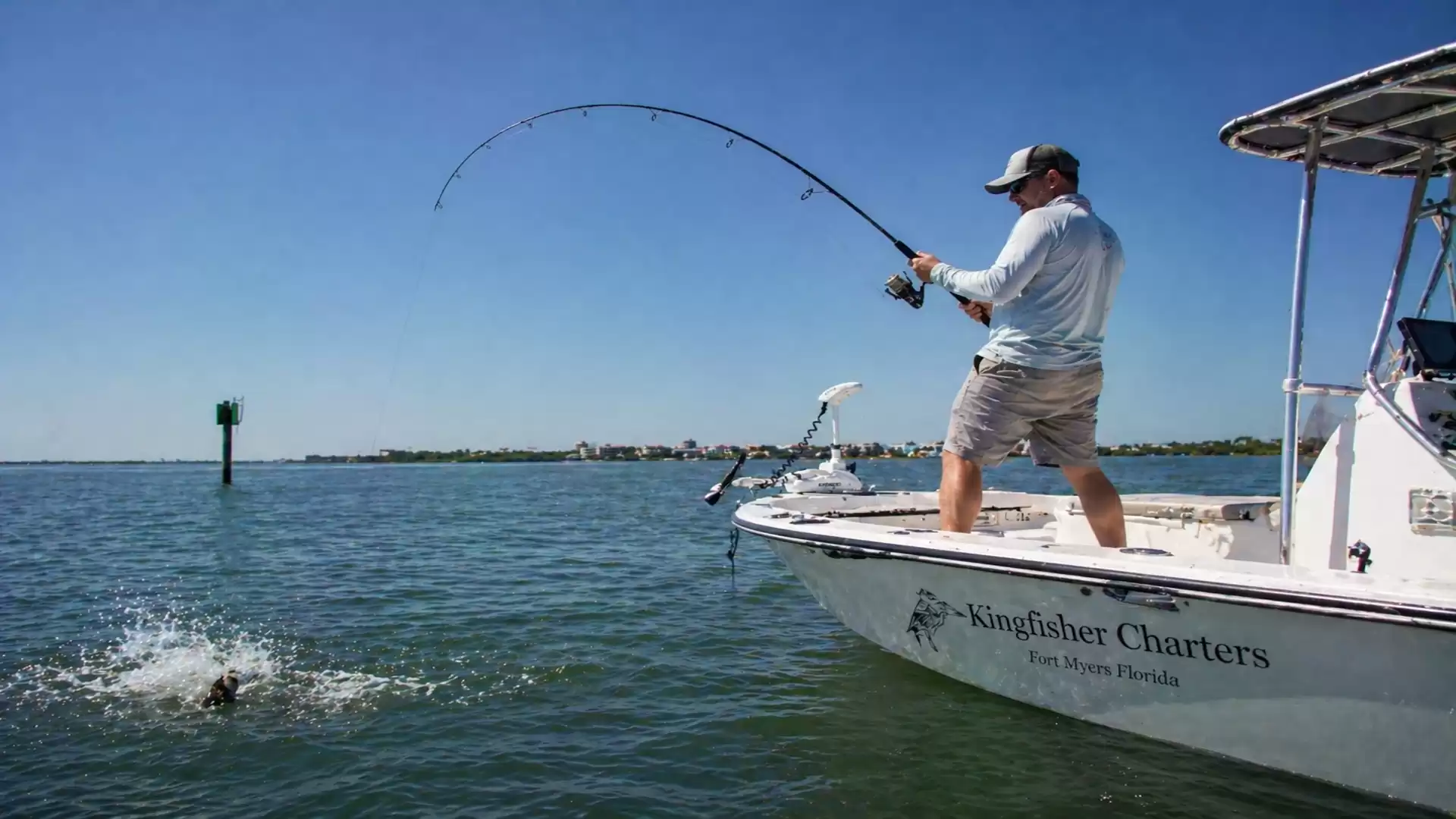 An angler stands on a boat and reels in a fish from the water while holding a fishing rod. The sky is clear and blue.