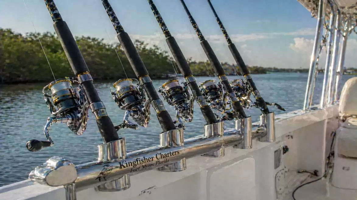 Six fishing rods in holders on a boat by the riverbank, surrounded by trees.