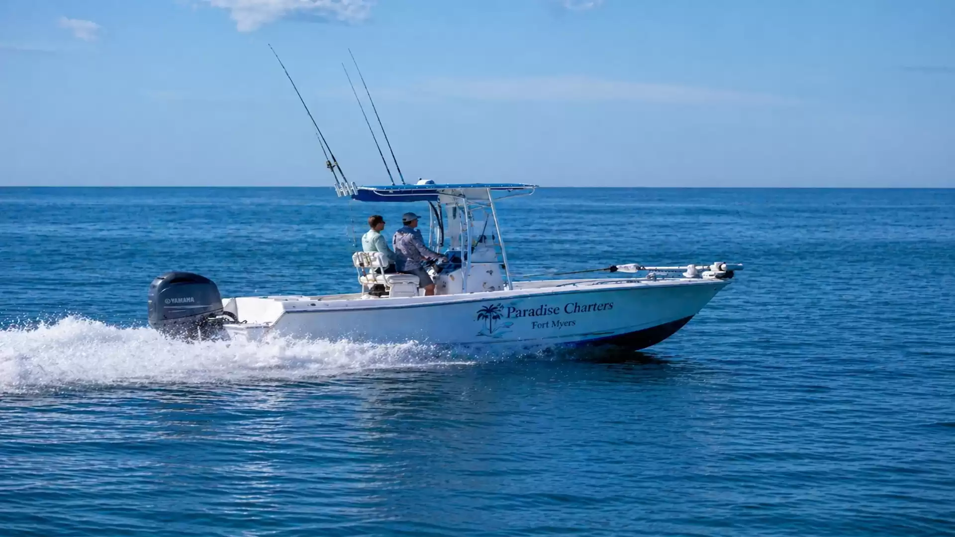 A boat sails on the blue ocean in sunny weather. Two people are on board steering the boat. The sky is clear with few clouds.