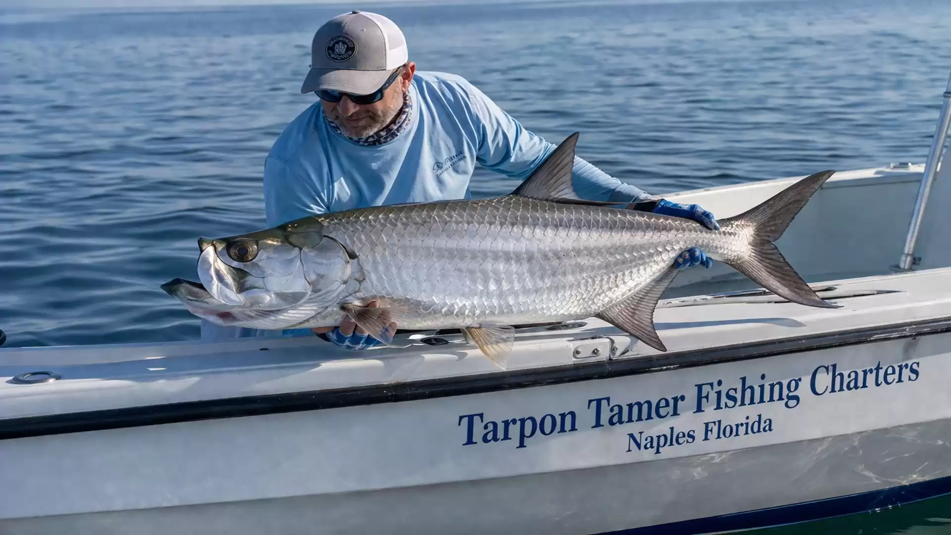 An angler on a boat holds a large tarpon fish and smiles. The boat reads 'Tarpon Tamer Fishing Charters Naples Florida'.