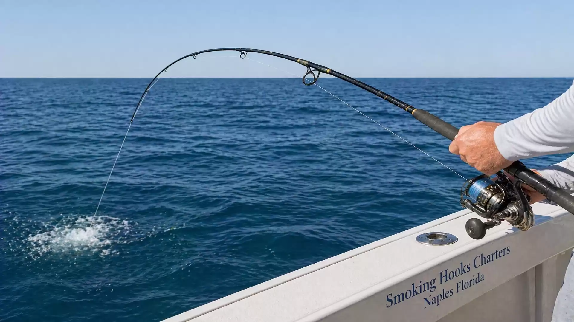 Angler on a boat struggles with a fish, while the fishing rod is bent. The ocean is calm and the sky is clear.