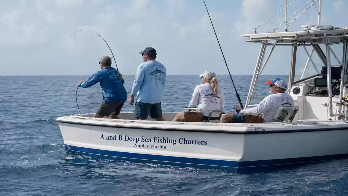 Four people deep sea fishing on a small charter boat in the open sea. One of the fishers has a fishing rod cast, while the others watch.