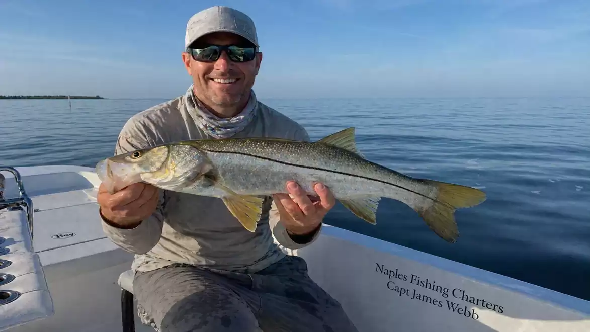 A smiling man on a boat holding a caught fish in front of a calm sea backdrop.