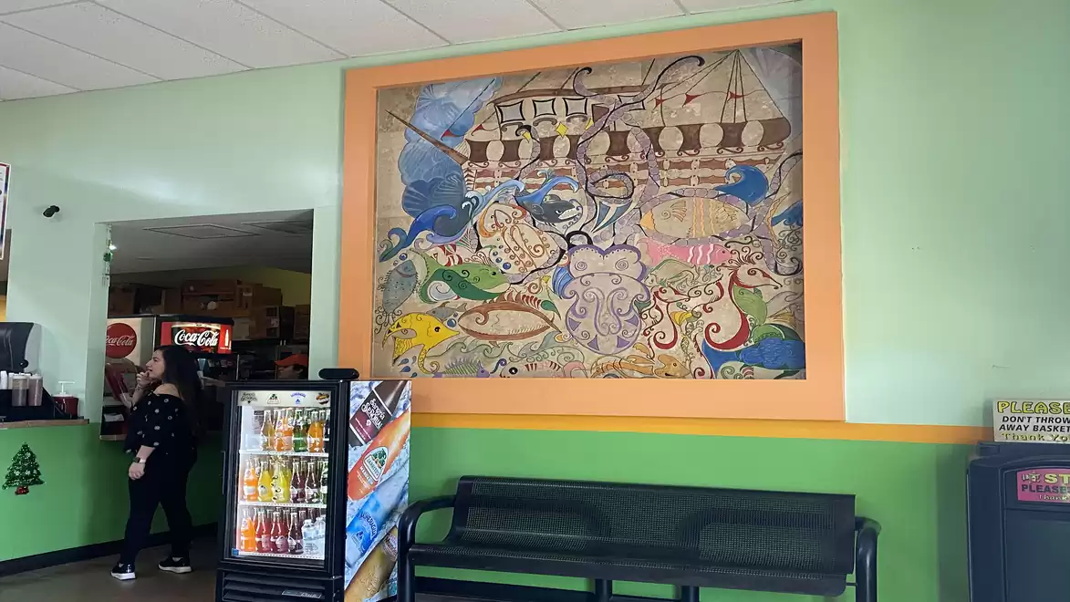 An indoor scene of a store with green walls, an artistic mural of a ship and sea scenes, a vending machine, a woman in the background, and a seating bench.