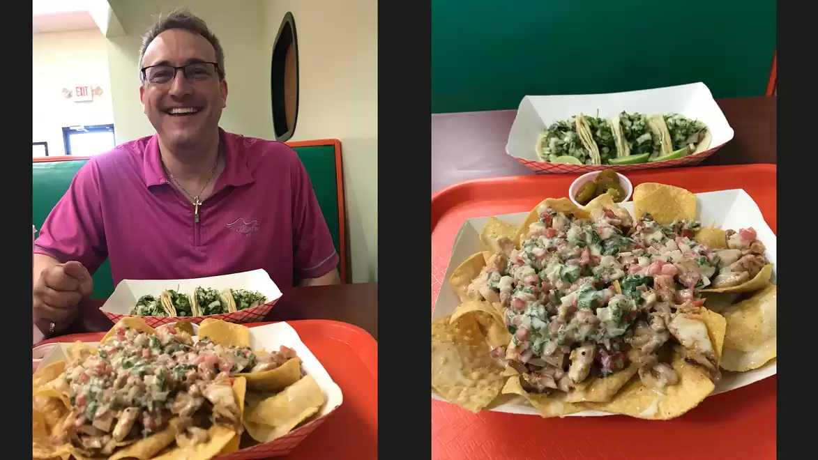 A smiling man in a pink shirt is sitting at a table in a restaurant. In front of him is a tray with nachos and small tacos.