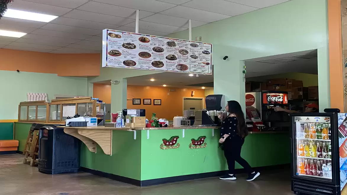 Interior of a small restaurant with a green counter, a person standing at the counter. Menu on the wall, drink dispenser and fridge visible.