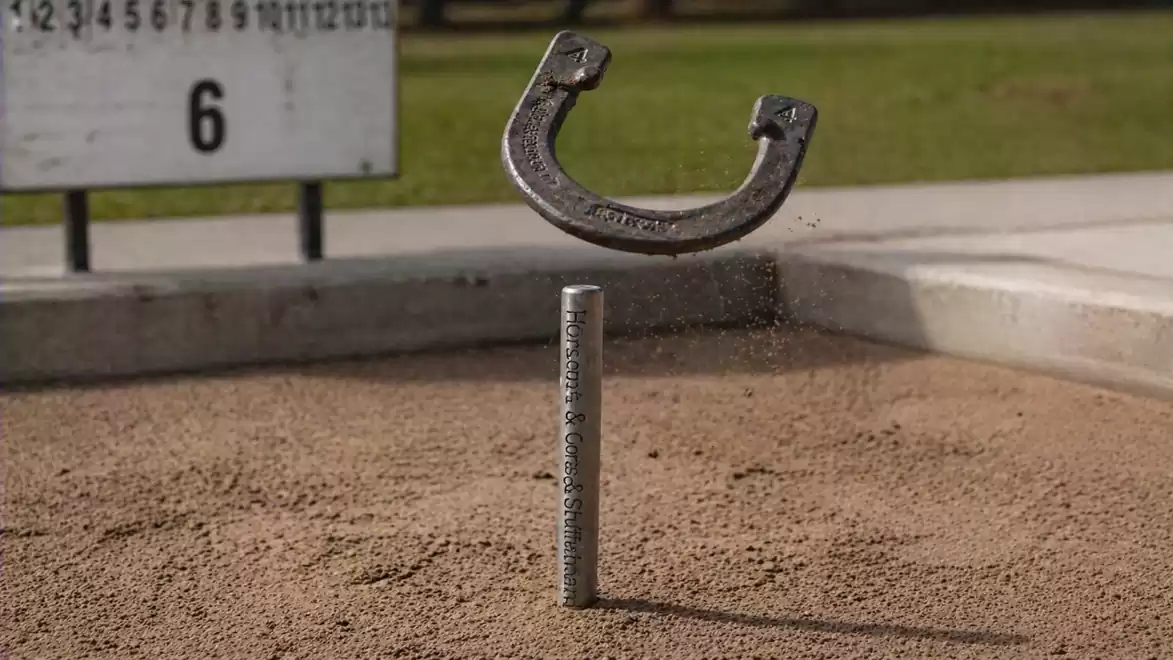 A horseshoe hovers in the air above a stake at a pitching facility, surrounded by a sand area.