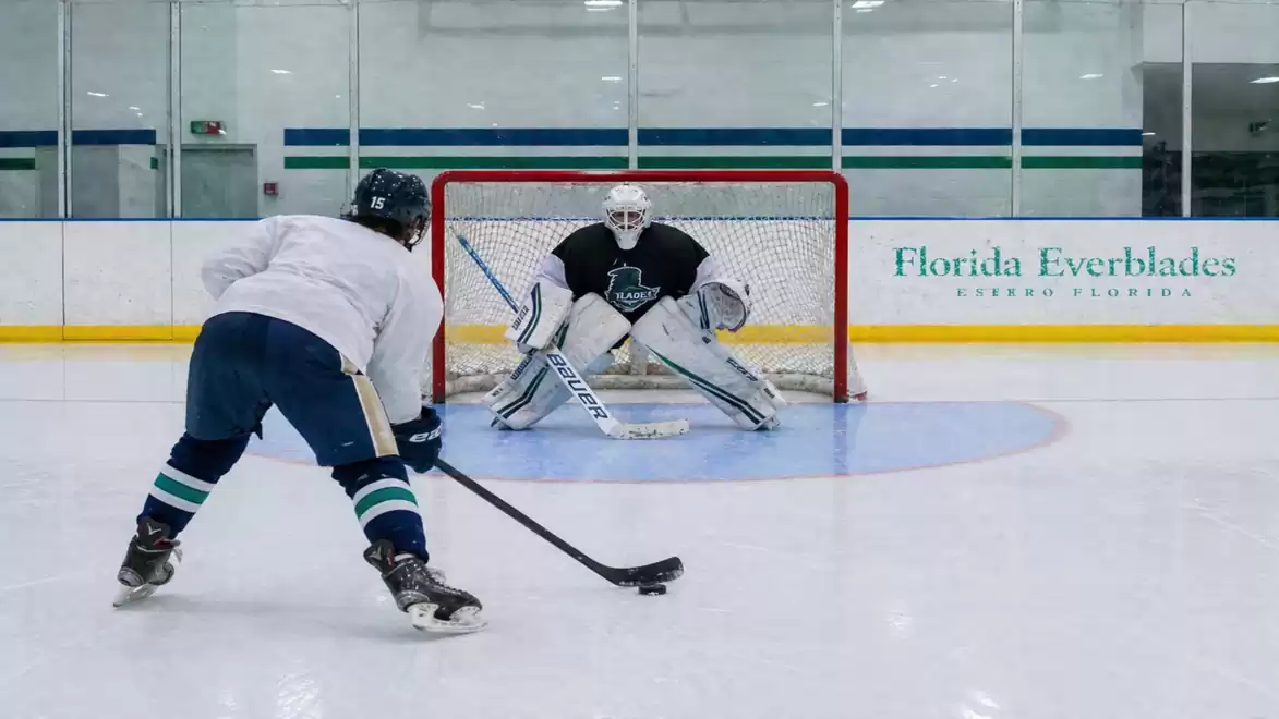 A hockey player in a white jersey faces a goalie on the ice, preparing to take a shot.