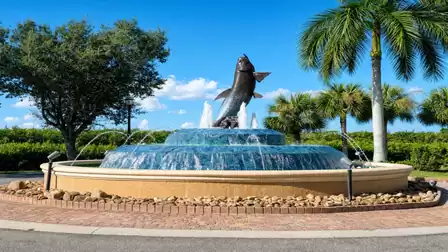 Round tiered fountain with leaping fish sculpture, surrounded by palm trees and shrubs