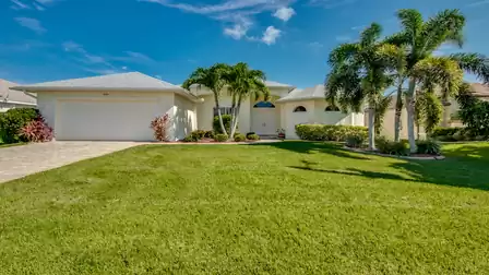 Elegant Villa Donatella in Cape Coral with a well-maintained front yard, palm trees, and tropical landscaping. Inviting entrance area with double doors and Mediterranean charm.