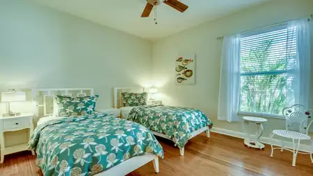 Bright bedroom with two single beds, turquoise floral bedspreads, white nightstands, and a large window overlooking greenery.