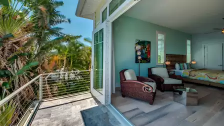 Open balcony door showing the bedroom, tropical plants, and stylish rattan furniture on the terrace.