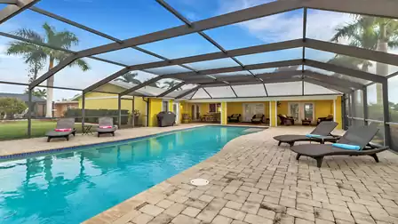 Covered pool area with seating group, sun loungers, and BBQ in front of yellow vacation home.