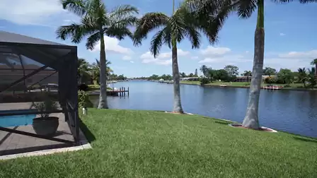View across the manicured lawn to a wide canal, with palm trees in the foreground and a pool area on the left.