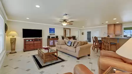 Living area with sofa, TV, ceiling fan, and view into the open kitchen with breakfast bar - a central spot for socializing and relaxation.