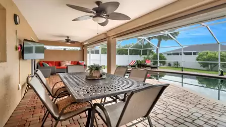 Dining area with a large table and six chairs under the roofed patio, facing the pool and enclosed by a breezy screen.