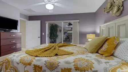 View from the bed toward the patio access, featuring a purple accent wall, ceiling fan, and cozy yellow-and-white bedding