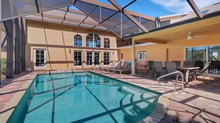 Covered patio with dining table and loungers at a sunny pool area with screen and tall windows in the background