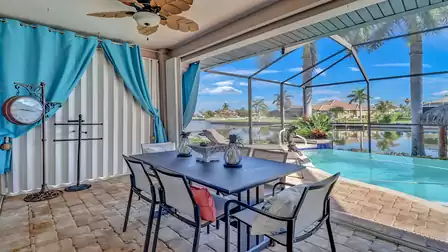 Covered outdoor dining area with a view of the pool and canal, framed by palm trees and turquoise curtains.