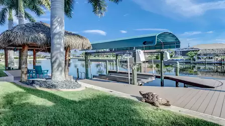 Boat dock with boat lift on the canal, in front a tiki hut with two blue Adirondack chairs.