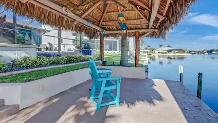 Cozy tiki hut right by the water with two turquoise high-back chairs and a ceiling fan.