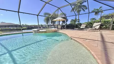 Spacious pool area with shallow entry, spa, sun deck and canal view surrounded by palm trees and tropical scenery.
