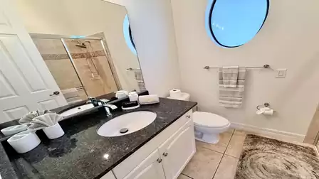 Bright bathroom with round window, granite vanity, glass-enclosed shower, and neutral tile finishes.