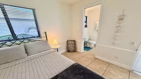 Bedroom with king size bed, nightstand and lamp; view toward open bathroom door, light tiles and calm tones.
