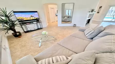 Bright living room with large sofa, glass coffee table, TV and open layout with views toward the pool area.