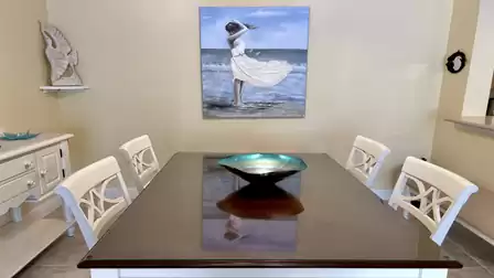 Wooden dining table with a glass top and four chairs, with a beach-themed wall artwork above.