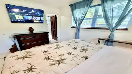 Bright bedroom with a comfortable double bed, smart TV, and large windows that fill the room with natural light. The tropical-inspired décor and peaceful ambiance create the perfect setting for a relaxing vacation.