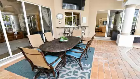 Covered poolside dining set on blue rug facing bar counter with wall mounted TV