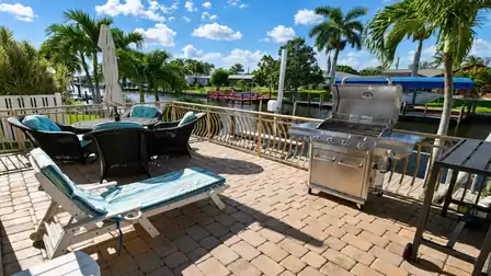 Canal terrace with wicker lounge chairs, striped sun lounger, gas barbecue and palm trees