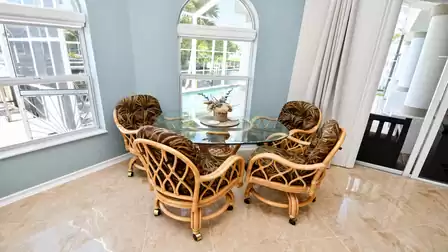 Dining corner with round glass table, four rattan chairs and windows toward pool area