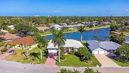 Aerial view of a quiet residential area with villas, palm trees and a lake in a sunny coastal setting.